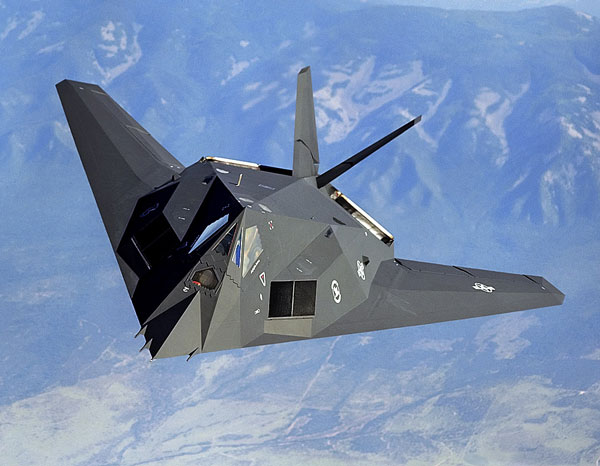 F-117 Nighthawk stealth fighter jet flying against a blue sky with mountainous landscape below.